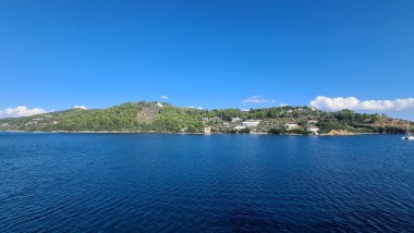 Uçsuz bucaksız mavi bir okyanusun manzarası. Arkasında gün ışığında yeşil dağlar var.
