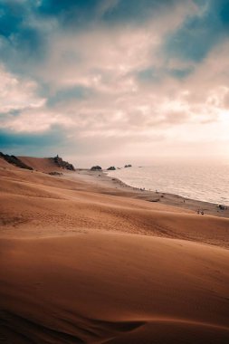 Kıyı şeridinin önündeki kumlu bir tepenin dikey görüntüsü ve arka planda muhteşem bulutlu bir gökyüzü.