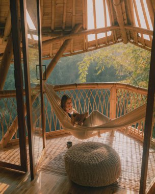 Arka planda ahşap bir çit ve yeşillikle hamakta yatan bir kadının dikey görüntüsü.