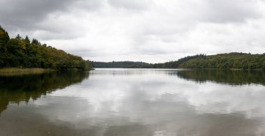 Sonbaharda, bulutlu bir günde, ormanlık bir sahilin yansımasıyla gölün yüzeyinin nefes kesici bir görüntüsü.