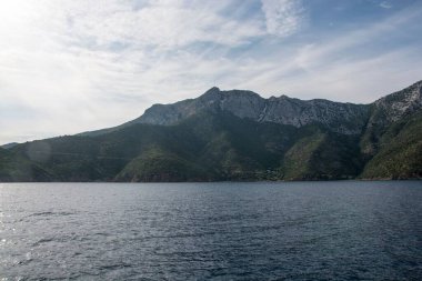 Bulutlu bir gökyüzüne karşı kayalık dağlarla çevrili büyüleyici bir göl manzarası.