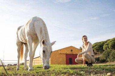 Genç ve mutlu bir binici kadın. Bir çiftliğin yakınında atıyla poz veriyor ve eğleniyor.