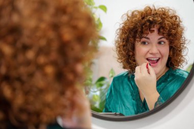 Banyo aynasına makyaj yapan kıvırcık saçlı tombul bir kadın.