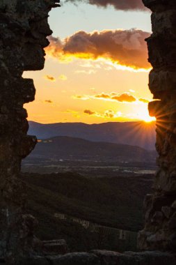 Günbatımı sahnesi. Işıklı, süzülen bulutlar yeşil bir manzaranın üzerinde. Kayalardan oluşan bir geçitten görülüyor.