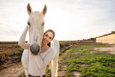 Genç ve mutlu bir binici kadın. Bir çiftliğin yakınında atıyla poz veriyor ve eğleniyor.
