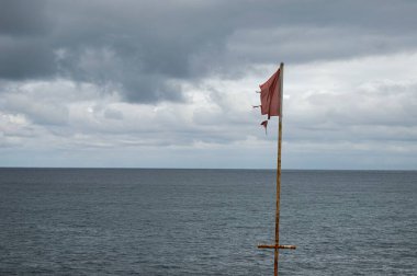 Bulutlu bir günde deniz kıyısında kırmızı dalgalı bir bayrak.