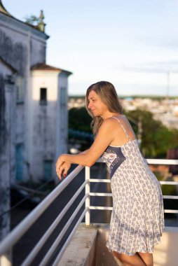 A woman on the porch of her house looking at the camera against the sky and church in the background