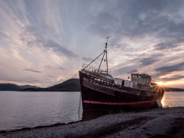 Eski terk edilmiş bir gemi Fort William sahilinde, bulutlu günbatımı gökyüzünün altında.