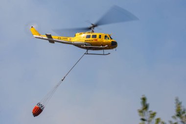 Huey helikopterinin, yangını söndürmek için bir kap su taşıyan alçak açılı görüntüsü.