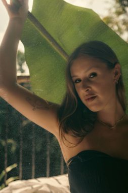 Siyah bir bluz içinde tropikal bir yaprağa tutunmuş beyaz bir erkeğin dikey görüntüsü.