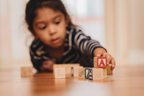 A selective focus shot of wooden letter blocks, with a preschool girl in the background, lying on the floor at home, the concept of early education