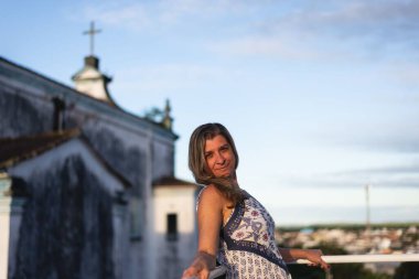 A woman on the porch of her house looking at the camera against the sky and church in the background