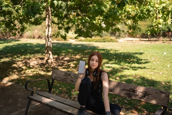 一个女孩坐在长椅上 展示着一个带有空白屏幕的手机 — 图库照片