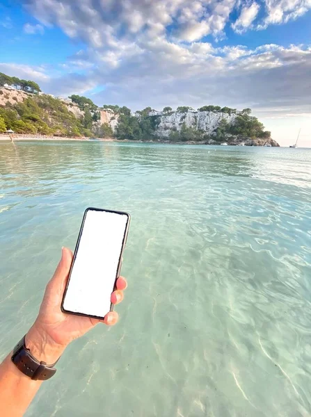 Deniz kıyısı seyahat uygulaması kavramının yakınında boş bir arkaplanı olan akıllı telefonu tutarken çekilmiş dikey bir fotoğraf.