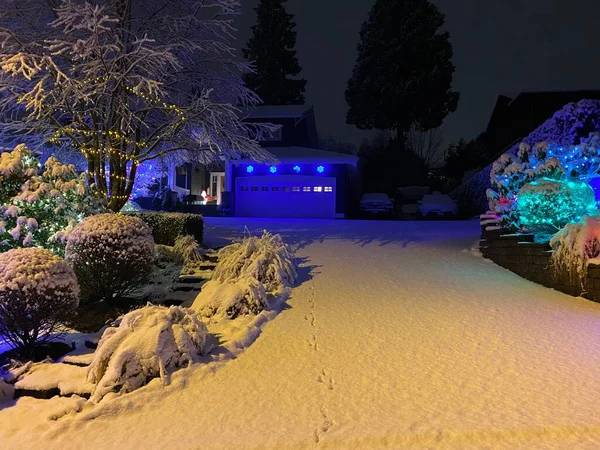 A snowy front garden and trees of a house in the background of other residual houses during the evening