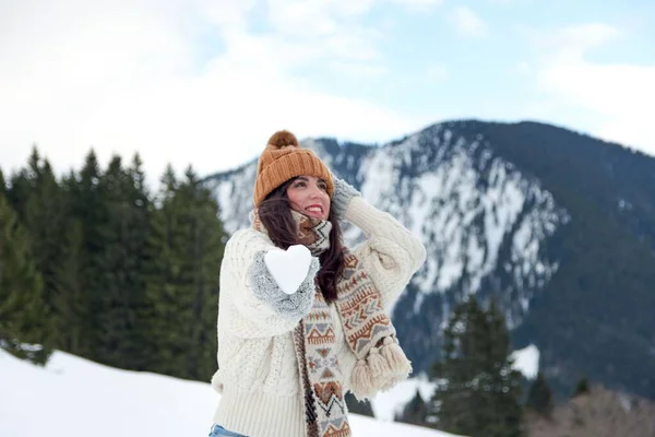 スカーフと冬の帽子をかぶった女性の観光客は カメラに向かってハート型の雪玉を保持します — ストック写真