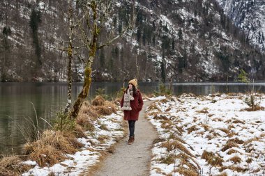 Almanya, Bavyera 'daki Konigssee Gölü' nde yürüyen kahverengi ceketli ve atkılı maceraperest bir kız.