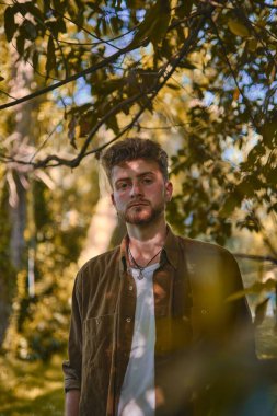 Üzerinde bitki örtüsü ve güneş ışığı olan ceketli sarışın bir erkeğin dikey portresi.