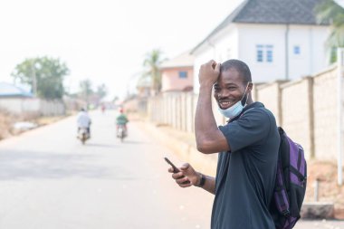 Yüzü maskeli Afrikalı bir adam dışarıda telefonunu kullanıyor.