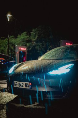 Geceleri yağmurun altında şarj olan Tesla arabasının dikey görüntüsü.