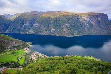Gökyüzü bulutlu arka planda, kırsal alanda büyük dağlarla çevrili mavi bir nehrin hava görüntüsü.