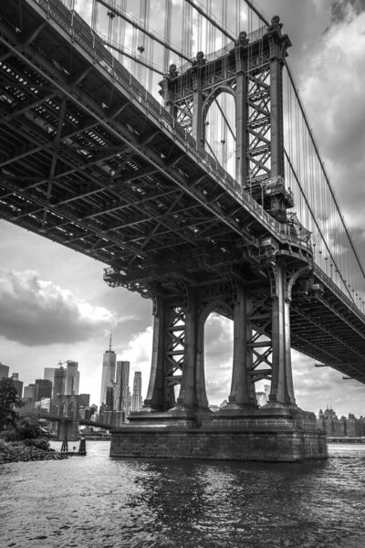 A low-angle shot of The Brooklyn Bridge over The East River in New York, with a cityscape in the background, in a grayscale