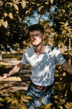 Ahşap bir çitin yanında, yemyeşil ağaçlarla çevrili beyaz bir adamın dikey görüntüsü.