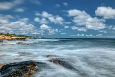 Beyaz dalgalı bir deniz manzarası ve bulutlu bir gökyüzüne karşı kayalık bir plaj.