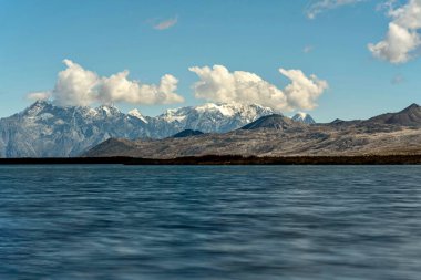 Gökyüzündeki kabarık bulutların altındaki dağlara karşı çekilen mavi bir deniz manzarası.