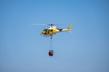 Mavi gökyüzünde suyla dolu bir sepet taşıyan itfaiye helikopteri.