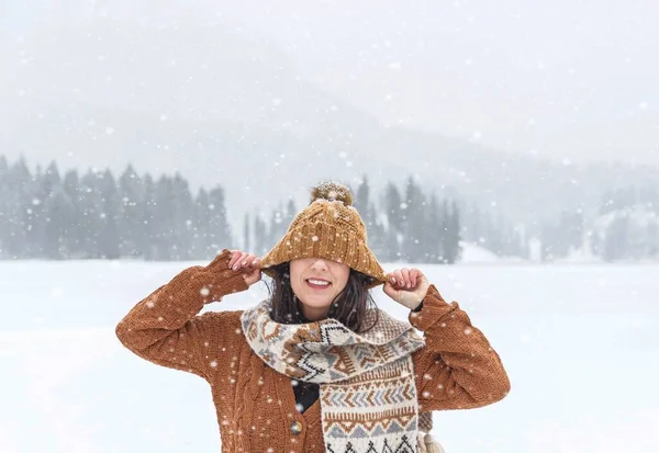 一个穿着舒适的冬季毛衣和围巾的女人把她的帽子蒙在眼睛上 在雾蒙蒙的冬季森林里微笑着 — 图库照片