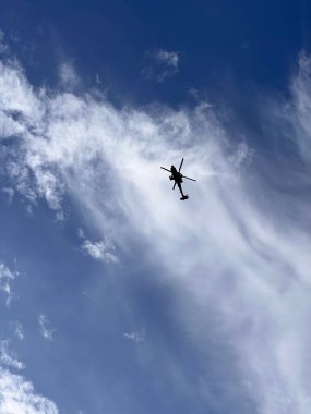 Mavi gökyüzünde beyaz bulutlarla uçan bir helikopter silueti.