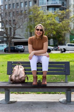 Halka açık bir parkta bankta oturan gülümseyen bir kadının dikey çekimi.