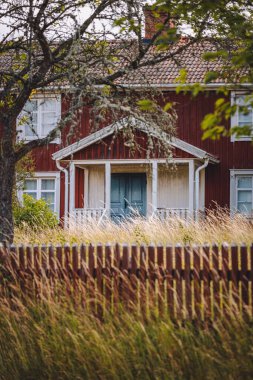 Ağaçlar ve otlarla çevrili eski kırmızı ahşap bir evin dikey görüntüsü.