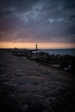 Güneş ufukta batarken deniz fenerinin dikey görüntüsü görünür.