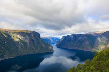 Gökyüzü bulutlu arka planda, kırsal alanda büyük dağlarla çevrili mavi bir nehrin hava görüntüsü.
