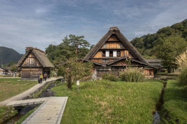 Bir grup turist, Japonya 'nın tarihi bir köyü olan Shirakawa-go ve Gokayama' yı ziyaret ediyor. Ağaçlarla çevrili, arka planda ormanlı bir dağ bulunuyor.