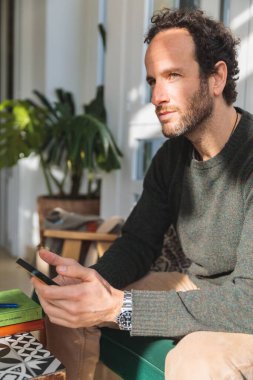 Kanepede oturan beyaz adamın dikey bir fotoğrafı. Elinde akıllı bir telefon var ve düşünüyor.