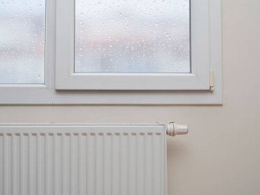 Pencerenin altında termostatı olan beyaz, modern bir radyatör.