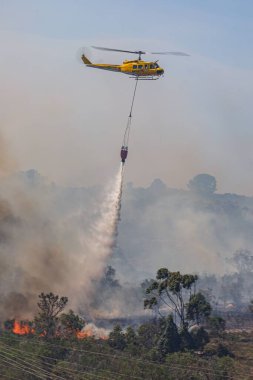 Huey helikopterinin alevler içindeki bir su torbasını düşürdüğü dikey bir görüntü.