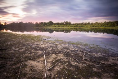 Gün batımında ormandaki ve gökyüzündeki ağaçları yansıtan berrak bir su kıyısı.