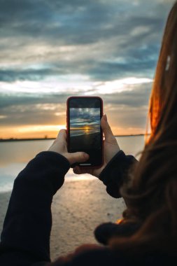 Gölde telefonuyla gün batımında fotoğraf çeken genç bir kadın.