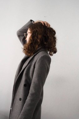 Kıvırcık kahverengi saçlı ve beyaz arka planda poz veren resmi olan genç bir kadının dikey çekimi.