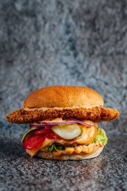 Gri bir yüzey üzerinde sebzeli büyük boy kızarmış tavuk çizburgerinin dikey yakın çekimi.