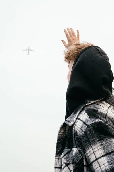 空を飛ぶ飛行機に手を振っている若い男性の垂直ショット — ストック写真