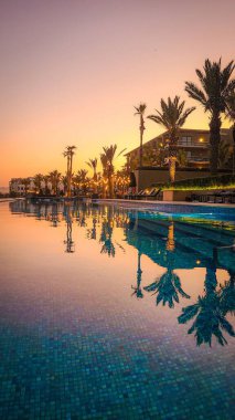 Tropikal tatil köyü üzerinde, palmiye ağaçları ve havuzu olan gün batımının dikey görüntüsü.