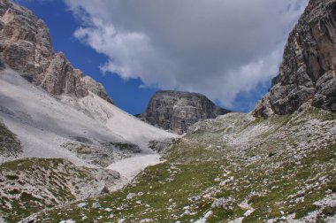 Tepenin yamacı, kısmen otla kaplı ve engebeli dağlarla çevrili, mavi gökyüzünde yoğun beyaz bir bulut.
