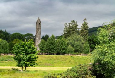 Yuvarlak Kule, bulutlu gökyüzünün arka planında yeşil ağaçlarla çevrili. Glendalough.