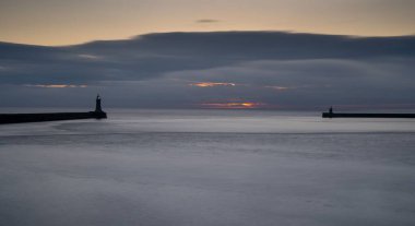 Gün doğumunda Tyne nehrinin ağzındaki sakin su, rıhtım ve deniz fenerlerinin manzarası.