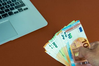 Sarı yüzeydeki bir dizüstü bilgisayarın yanında euro banknotlarını tutan bir el, online ticaret kavramı